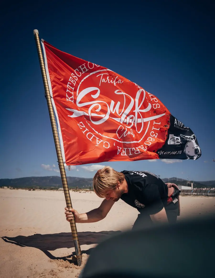 Monitor de Tarifa Surf Club clavando la bandera roja del club en la playa de Tarifa